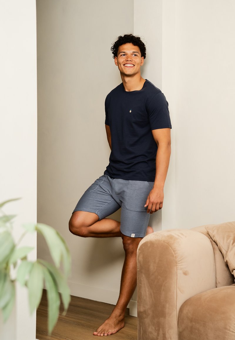 Navy t-shirt met een klein embleem, gecombineerd met lichtgrijze geblokte shorts. Model staat blootsvoets tegen een muur, met een pluche bank in de buurt.