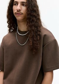 Tricou oversized maro, realizat din bumbac moale, cu mâneci scurte, complementat de coliere suprapuse, incluzând o lanț din argint și un fir cu perle.