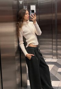 Maglione bianco a coste con zip, maniche lunghe e colletto alto, abbinato a pantaloni neri larghi. Sfondo di un ascensore metallico liscio.