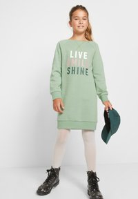 Ragazza con coda di cavallo che indossa un vestito verde con la scritta "LIVE SMILE SHINE", collant bianchi, stivali neri a fiori, mentre tiene un cappellino da baseball verde scuro.