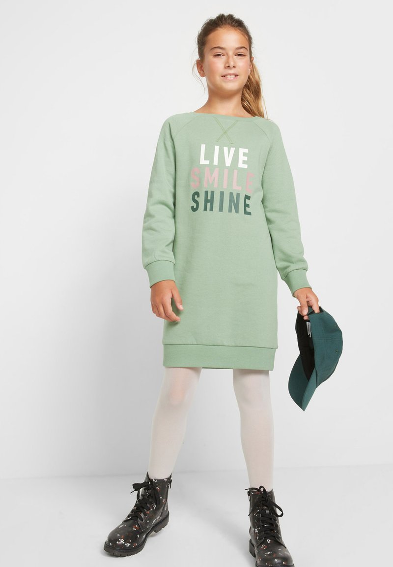 Ragazza con coda di cavallo che indossa un vestito verde con la scritta "LIVE SMILE SHINE", collant bianchi, stivali neri a fiori, mentre tiene un cappellino da baseball verde scuro.
