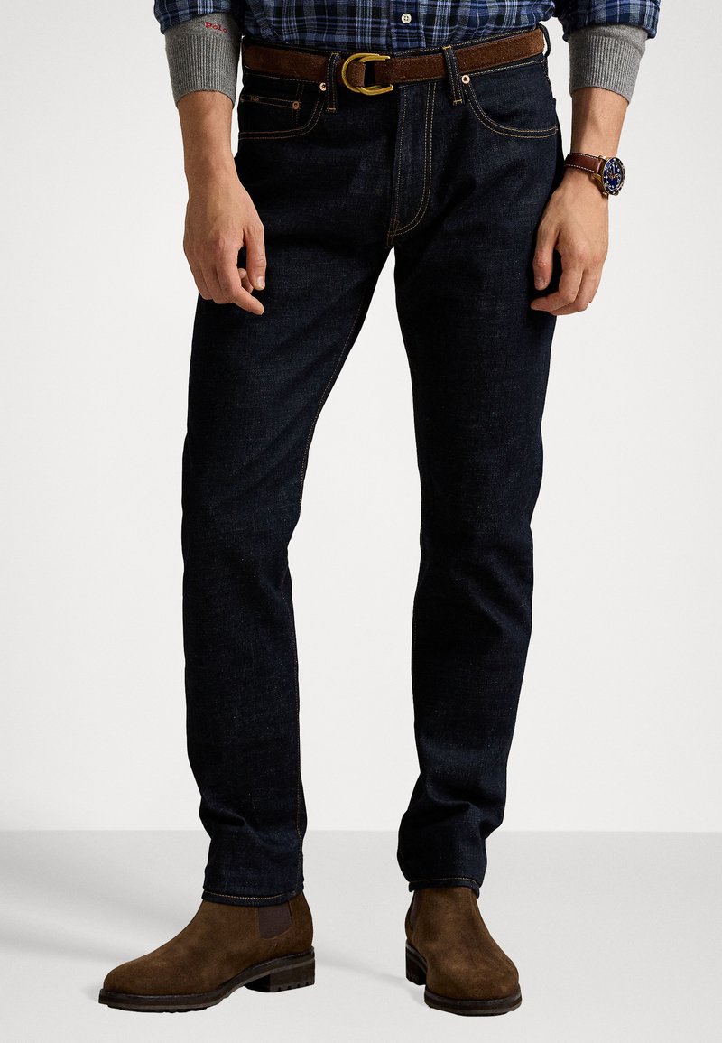 Jeans in denim blu scuro con una vestibilità slim, caratterizzati da cuciture a contrasto e passanti per cintura. Indossati con una cintura in pelle marrone e stivali in camoscio marrone.