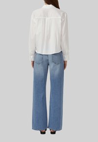Camisa blanca de manga larga con cuello abotonado, combinada con unos vaqueros de piernas anchas en un tono azul claro que presentan bolsillos traseros y un efecto deslavado.