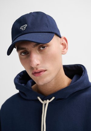 Navy blue baseball cap with a bee emblem on the front, made of cotton twill, paired with a navy hoodie featuring a cream drawstring.