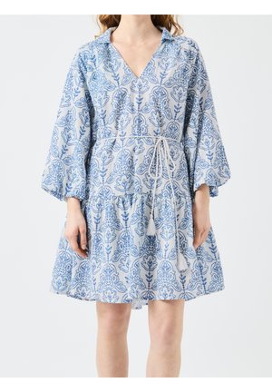 Femme portant une robe bleu clair avec des motifs floraux bleu foncé, manches longues bouffantes, encolure en V, et une ceinture à nouer avec des pampilles.