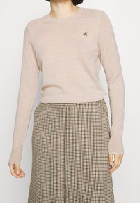 Pull en maille rose clair avec des manches longues et un détail côtelé à l'épaule, featuring un petit motif floral brodé sur la poitrine.