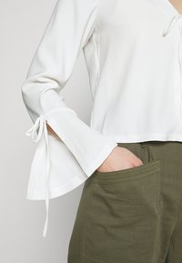 Blusa bianca con maniche a campana, caratterizzata da decorazioni con lacci ai polsini. Abbinata a pantaloni verde oliva con tasca laterale. Texture morbida.