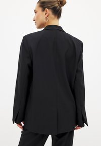 Črn krojen blazer z dolgim, ravnim krojem, mehko teksturo in ozkim zareznim ovratnikom, z razporkom na hrbtu za lažje gibanje.