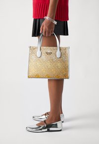 Gold and silver patterned handbag with two light gray handles, held by a person wearing a red sweater and black skirt, with silver flats.