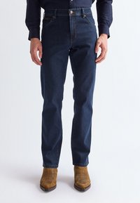 Homme portant un jean bleu foncé, des bottes en daim marron et une chemise sombre à manches longues, debout devant un fond uni.