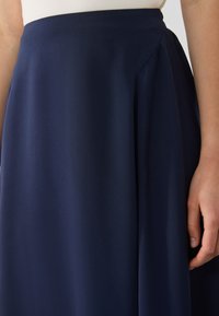 Gonna a pieghe blu navy in un tessuto fluido, caratterizzata da una texture liscia e una vita aderente, con una mano appoggiata sul tessuto.