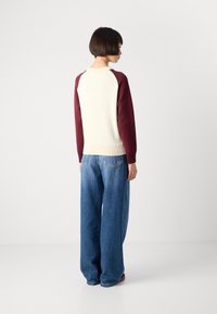 Person with short dark hair stands facing away, wearing a cream and burgundy long-sleeve top and wide-leg blue jeans against a plain background.