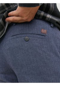 Tmavě modré kalhoty s rybí kostí vzorem, zadní kapsa se zapínáním na knoflík a malou hnědou koženou štítkem. Nošeny s kostkovanou košilí.