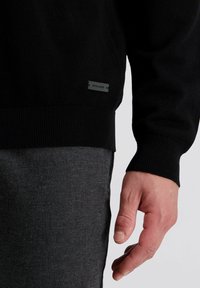Zwarte gebreide trui met geribde manchetten en zoom. Opvallend label met het "pierre cardin" logo aan de zijkant. Draagt over grijze met patronen bedrukte broek.