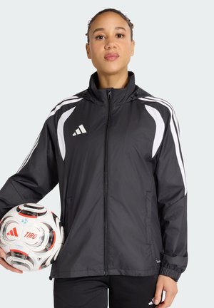 Person trägt eine schwarze Adidas-Jacke mit weißen Streifen und hält einen weißen und schwarzen Adidas Tiro Fußball auf Taillenhöhe.