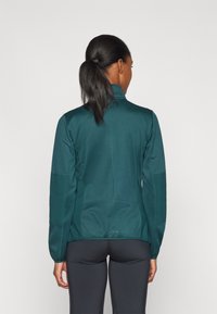 Giacca sportiva teal con colletto alto, maniche lunghe e design del retro senza cuciture. Pannelli testurizzati ai lati aggiungono dettagli al tessuto.