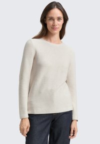 Svetre svetle bež barve z rebrastim vzorcem, dolgimi rokavi in okroglim izrezom, kombiniran z temno modrimi jeans hlačami. Mehka tekstura in sproščen kroj.