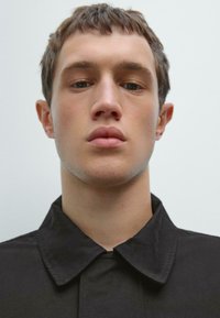 Black collared shirt with a smooth texture, featuring a concealed front placket, model displays a neutral expression against a light backdrop.
