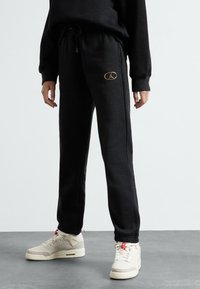 Crne jogger hlače napravljene od mekanog materijala, s podesivim pojasom, bočnim džepovima i zlatnim logotipom na lijevom bedru.