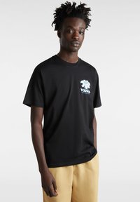 Camiseta de algodón negra con un gráfico de una palmera y el texto "VANS" en azul claro. Cuello redondo clásico, mangas cortas y corte relajado.