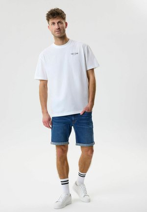 Uomo con t-shirt bianca "Gin & Tonic", pantaloncini di jeans blu risvoltati, calzini bianchi con strisce nere e sneakers bianche, in piedi su sfondo bianco.