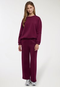 Completo composto da felpa bordeaux e pantaloni a gamba larga, realizzato in tessuto morbido. Presenta un girocollo e una vita elasticizzata, abbinato a sneakers argento.