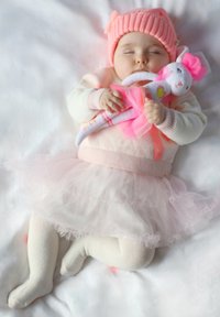 Neonato addormentato su una superficie bianca e morbida, indossando un cappello e un maglione rosa, con un giocattolo che ha una gonna e un fiocco, circondato da tessuti delicati.