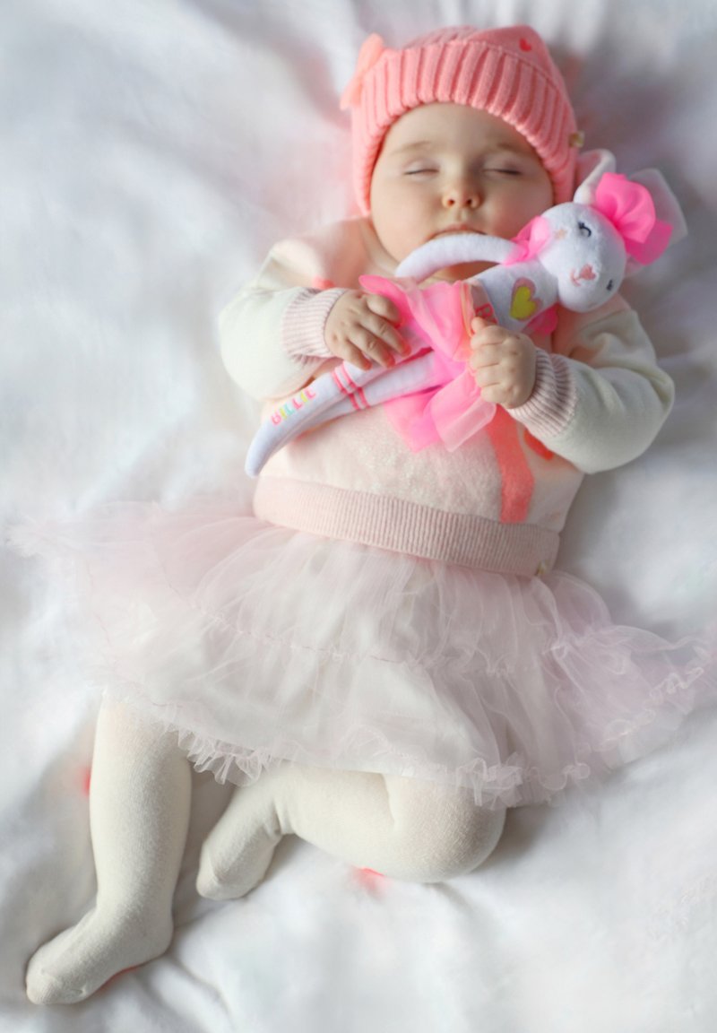 Neonato addormentato su una superficie bianca e morbida, indossando un cappello e un maglione rosa, con un giocattolo che ha una gonna e un fiocco, circondato da tessuti delicati.