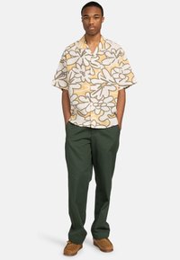 Joven con una camisa de manga corta holgada de flores amarillas y blancas, pantalones verde oscuro y zapatos de ante marrones, de pie con las manos en los bolsillos.