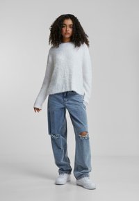Maglione bianco in maglia fuzzy con orlo leggermente corto, abbinato a jeans strappati di un blu chiaro. La modella indossa sneakers bianche. Sfondo semplice.