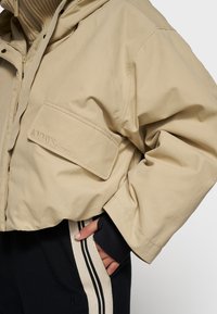 Beige cropped jakke med hætte, med en frontlomme og broderet logo. Båret over sorte bukser med kontraststriber på siden.