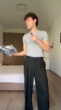 Jaunas vyras dėvi pilką marškinėlį ir juodas kelnes, laiko drabužį ir priežiūros šepetį šviesiame miegamajame su medinėmis grindimis ir spinta.