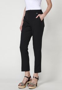 Pantalons ajustés noirs avec une texture lisse, dotés d'un devant plat, de poches latérales et d'une coupe slim, associés à des sandales compensées rayées.