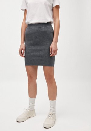 Femme portant un t-shirt blanc, une mini-jupe en tricot gris, des chaussettes blanches et des baskets blanches, debout devant un fond uni.