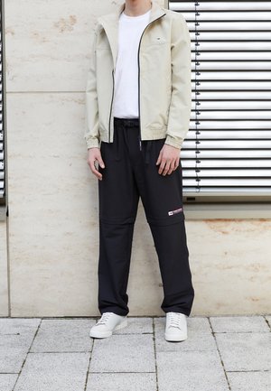 Personne debout portant une veste beige zippée, une chemise blanche, un pantalon noir avec un patch latéral et des baskets blanches contre un mur avec des stores de fenêtre.
