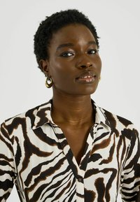 Blusa blanca con un patrón de rayas de cebra en negro y marrón, con cuello con solapa y cierre de botones. Pendientes de aro dorados.