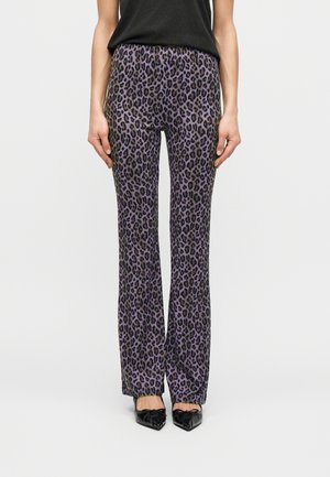 Femme portant un pantalon droit à imprimé léopard violet et des ballerines noires à bouts pointus avec un nœud, debout devant un fond uni.