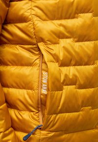 Giacca imbottita gialla con texture trapuntata, dotata di una tasca con zip etichettata "STOW AWAY" e una zip arancione a contrasto.