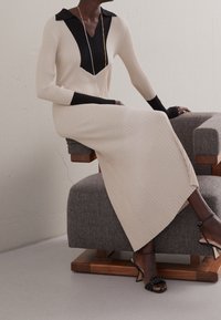 Abito in maglia con scollo a V nero e colletto, maniche lunghe a costine e corpo color crema. Indossato con sandali neri con cinturini.
