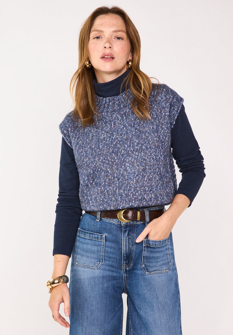 Un col roulé marine superposé sous un haut sans manches en tricot à pois bleus, associé à un jean taille haute et une ceinture marron.