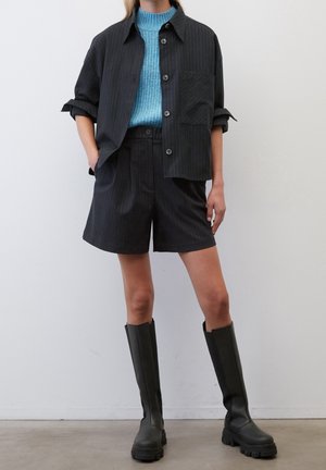 Czarna koszula w paski z kieszonką na piersi, niebieski sweter o fakturze na dole, czarne spersonalizowane szorty oraz czarne gumowe buty do kolan.