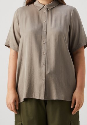 Vrouw draagt een losse, lichtbruine blouse met korte mouwen, knopen en kraag, en olijfgroene broek.