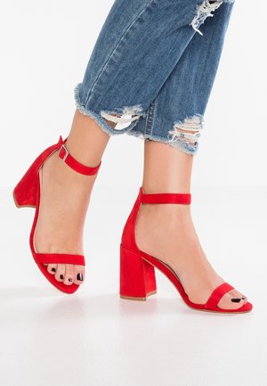 Czerwone zamszowe sandały na obcasie z masywnym, blokowym słupkiem i paskiem wokół kostki z klamrą. Otwarty nosek odsłania zadbane paznokcie i stopę.
