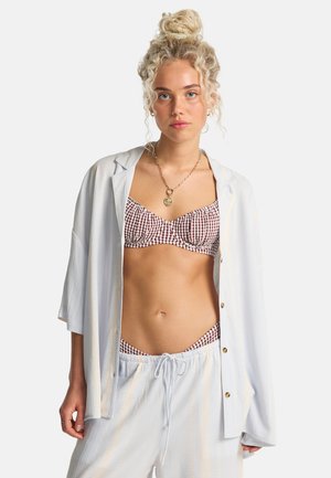 Blondynka z upiętymi włosami nosi czerwono-biały bikini w kratę pod luźną białą koszulą na guziki oraz pasujące spodnie z wiązanym paskiem, stoi przodem.