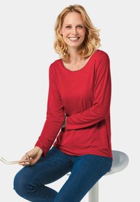 Rotes Langarmshirt mit weitem Ausschnitt, glattem Stoff und seitlichen Schlitzen, kombiniert mit blauen Jeans und in sitzender Pose mit einer Brille.