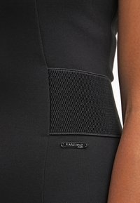 Close-up of black garment with wide textured waistband and small metallic "Marciano" brand tag near the hip on a person's side.