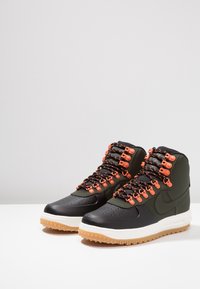 High-top sneakers made of black leather and green synthetic material. Features orange eyelets and laces, with a rubber gum sole.