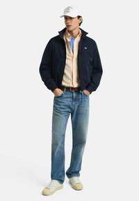 Marine bomberjack over een gestreept overhemd met knopen, blauwe jeans en lichte sneakers. De jas heeft een ritssluiting en geribbelde manchetten.