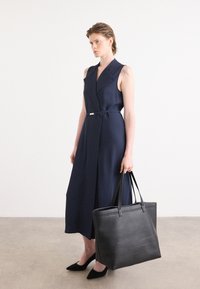 Vestido sin mangas de color marino con cintura ceñida, tejido texturizado, escote en V profundo y un bolso de cuero negro. La modelo lleva unos zapatos planos negros de punta.