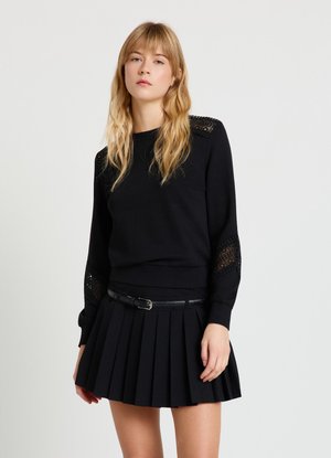 Jeune femme aux longs cheveux blonds porte un haut noir à manches longues avec des détails en dentelle et une jupe plissée noire avec une ceinture.